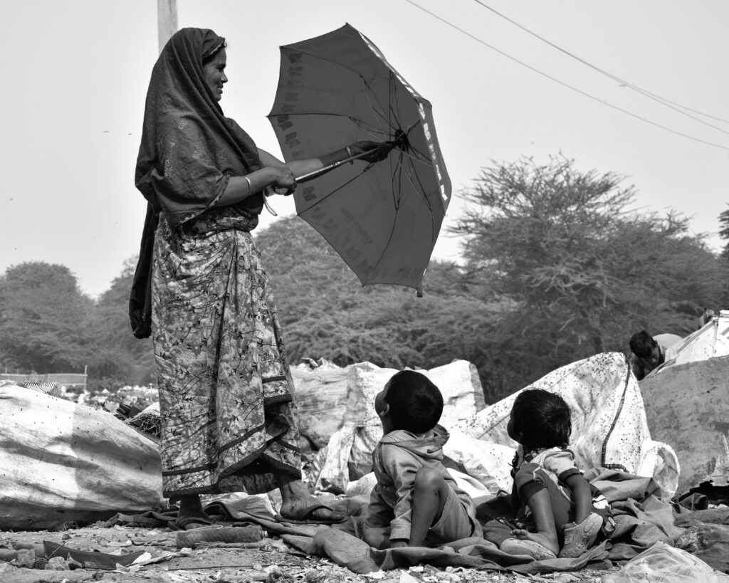 A working mother who is a Waste Worker opening an umbrella to protect her children from the Sun, women bring their children along with them to the waste segregation site which their workplace.